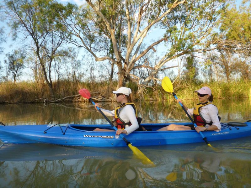 sunsmart young ladies paddle double kayak through narrow creek