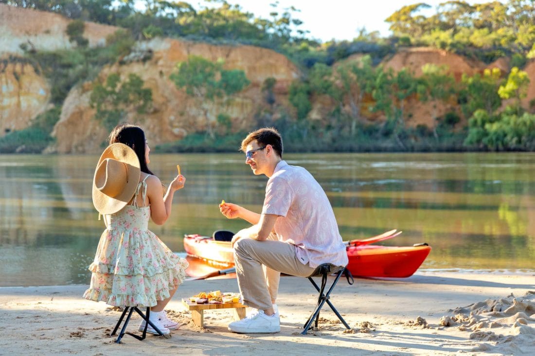 Couple seated on the riverbank, near a kayak, eating from a picnic platter.