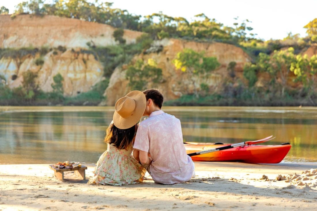 Couple kiss, sitting on the sand. Cliffs, River and kayak in background.