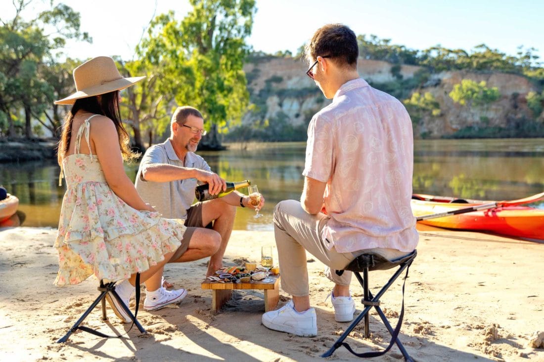 A couple sit at a low picnic table on a sandbar, with kayaks and cliffs in the background. A tour guide pours a glass of sparkling wine.