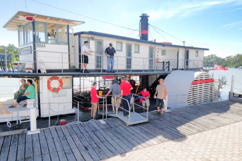 Passengers board a paddle steamer, via a short gangplank. The smoke stack is visible at the top of the boat, and the paddle wheel housing is visible on the boats side.