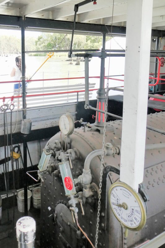 A black boiler housed in the centre of a paddle steamer, with rivets showing, as well as a steam gauge, and aa dial showing speed and direction of paddle wheels. 