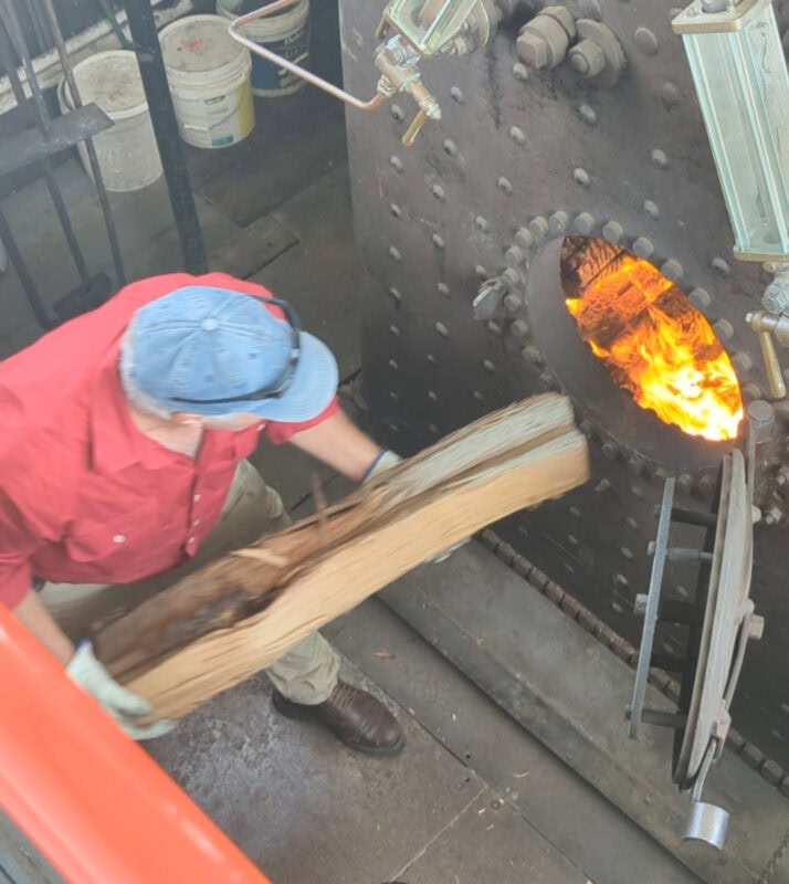 On a paddle steamer, looking at the boiler. The firebox door is open and a red-shirted man is putting a long piece of branch into the opening. Fire is visible inside the door.