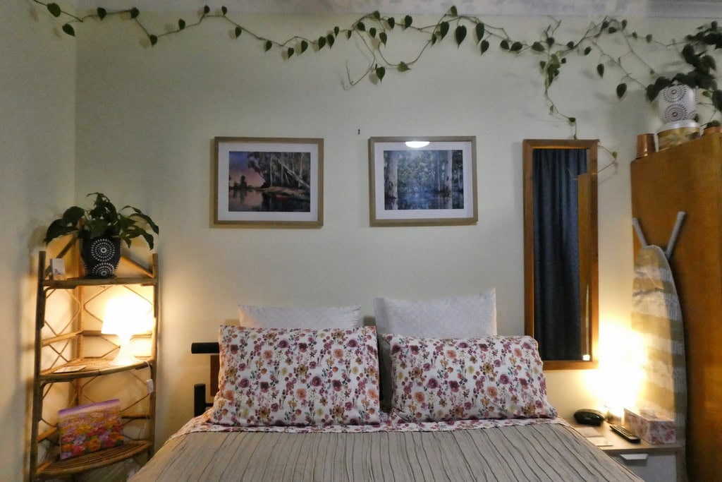Comfortable bed in Bed and Breakfast, with floral pillows, European pillows, bedside tables with glowing lamps, local artwork, plants and mirror.